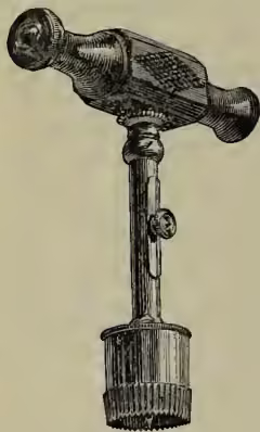 A detailed black and white illustration of a trephine, an ancient surgical instrument. It features a long, cylindrical handle with a textured grip, topped by a circular head with a central pin. The base of the handle is flared and has a small circular opening.