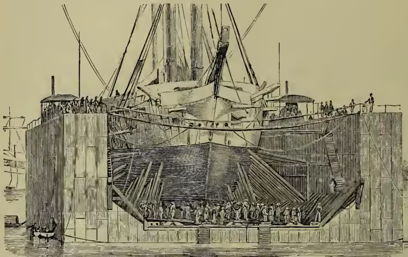 A detailed black and white illustration of a floating dock at Saigon. The dock is a large, rectangular structure made of vertical wooden planks, forming a box-like enclosure. It is shown partially submerged in water. A large ship with multiple masts and rigging is positioned inside the dock, its hull resting on the bottom of the structure. Several figures of people are visible on the deck of the ship and on the top of the dock structure. The water is calm, and the overall scene depicts the operation of a floating dock for maritime transport.
