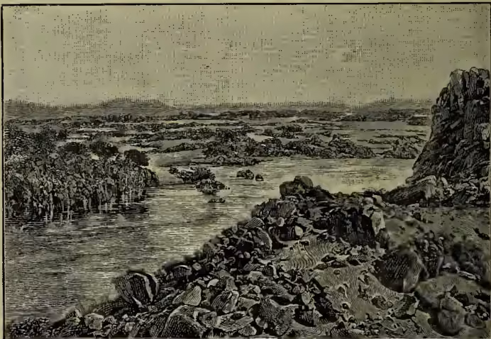 A detailed black and white engraving of the Nile river at the First Cataract. The river flows from the left towards the right, surrounded by steep, rocky cliffs. The water is turbulent as it passes through the narrow channel between the rocks. In the background, more distant hills and a clear sky are visible.