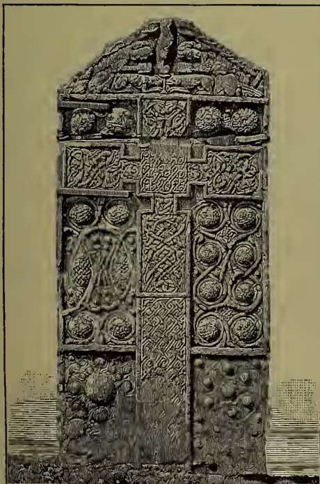 A detailed black and white photograph of a tall, rectangular sculptured stone. The stone is covered in intricate carvings, including a large central panel with a cross-like motif, surrounded by smaller panels with various figures and patterns. The top of the stone is decorated with a series of small, rounded carvings.